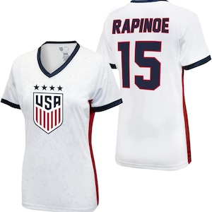 May include: White soccer jersey with red and navy blue accents. The jersey has the USA crest on the front and the name "RAPINOE" and the number "15" on the back.