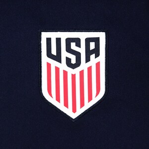 May include: A navy blue fabric with a white and red USA emblem. The emblem is shield-shaped with the letters "USA" in bold, white font at the top, and red vertical stripes below.