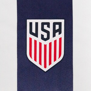 May include: Close-up of a white and navy blue striped fabric. A USA emblem is centered on the navy blue stripe. The emblem is a shield shape with the letters "USA" in navy blue and red vertical stripes.