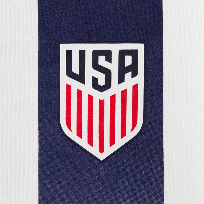 May include: Close-up of a white and navy blue striped fabric. A USA emblem is centered on the navy blue stripe. The emblem is a shield shape with the letters "USA" in navy blue and red vertical stripes.