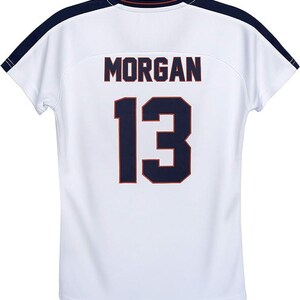 May include: White athletic jersey with the name "MORGAN" and the number "13" in navy blue with orange trim. The jersey has navy blue shoulder accents and a navy blue and orange trim on the collar.