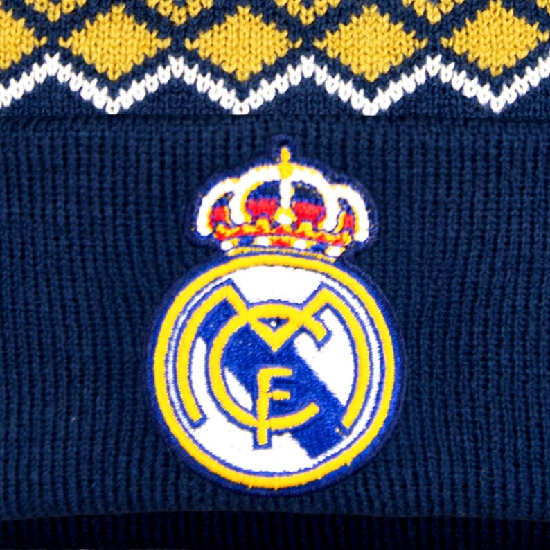 Puede incluir: Un gorro de punto azul marino con un patr&oacute;n de diamantes amarillos y blancos en la parte superior. El gorro tiene un parche circular con una corona y el logo del Real Madrid en amarillo, azul y blanco.