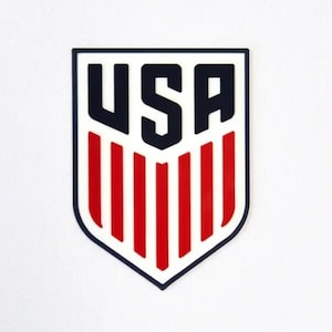 Puede incluir: Un logotipo azul y blanco en forma de escudo con la palabra "USA" en letras negras en negrita y rayas rojas y blancas.