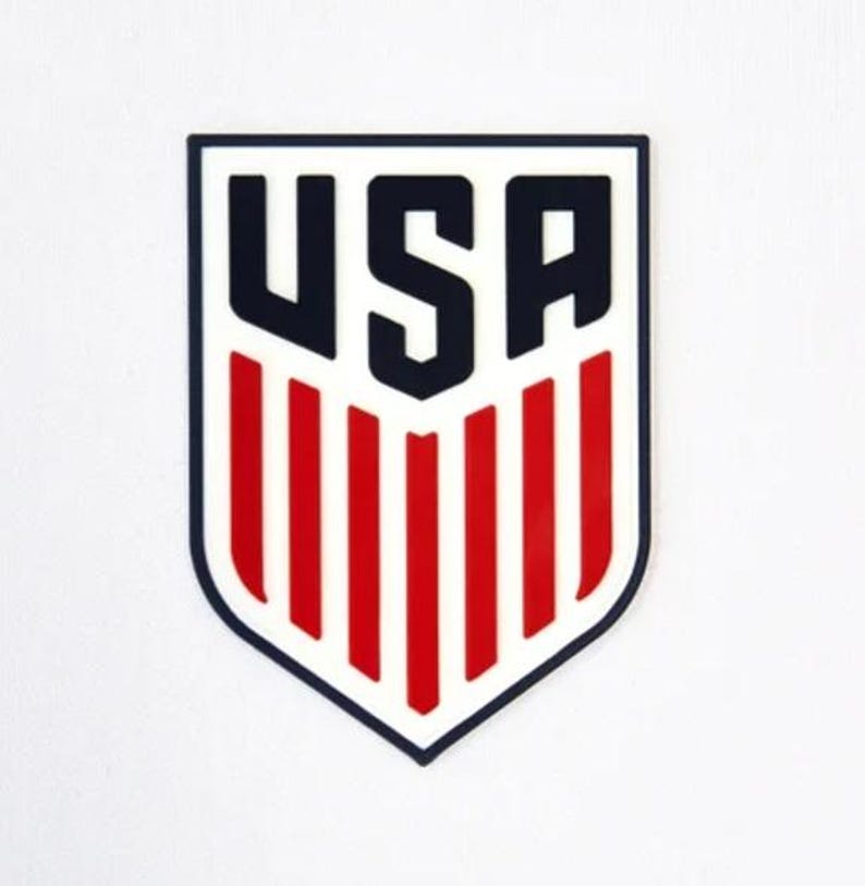 Puede incluir: Un logotipo azul y blanco en forma de escudo con la palabra "USA" en letras negras en negrita y rayas rojas y blancas.