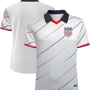 May include: White football shirt with a navy collar and red trim. The shirt has diagonal stripes in red, blue, and black. A USA crest is on the chest. The back of the shirt is plain white.