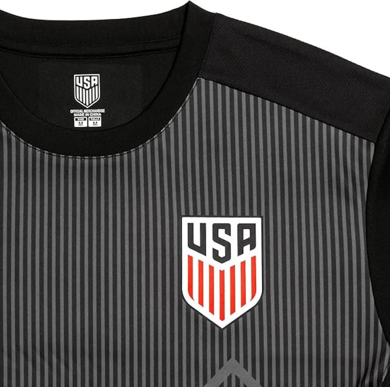 Puede incluir: Camiseta de f&uacute;tbol a rayas negras y grises con un logotipo de EE. UU. blanco y rojo en la parte delantera. La camiseta tiene un cuello negro y mangas cortas. El texto "USA OFFICIAL MERCHANDISE MADE IN CHINA" est&aacute; impreso en una etiqueta blanca unida al cuello.