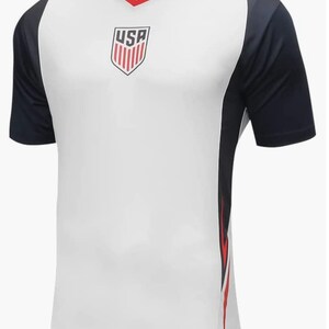 May include: White soccer jersey with a navy blue and red design. The jersey has a V-neck and the word "USA" is printed on the front.