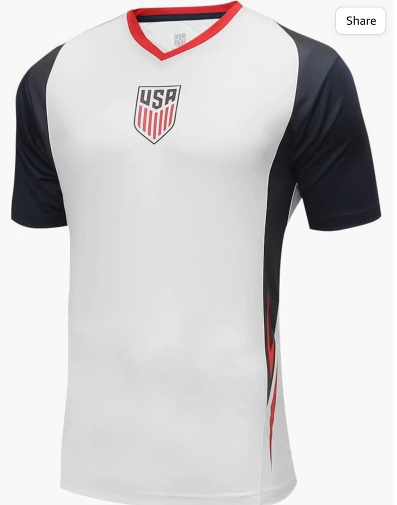 May include: White soccer jersey with a navy blue and red design. The jersey has a V-neck and the word "USA" is printed on the front.