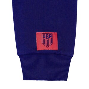 Op de afbeelding: Een marineblauwe trui met een rode patch op de mouw. De patch heeft een witte omtrek en het woord "USA" in blauwe letters. De patch heeft ook een gestileerd schild met rode en witte strepen.