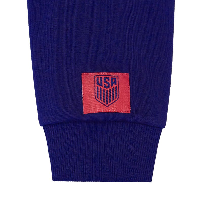 Op de afbeelding: Een marineblauwe trui met een rode patch op de mouw. De patch heeft een witte omtrek en het woord "USA" in blauwe letters. De patch heeft ook een gestileerd schild met rode en witte strepen.