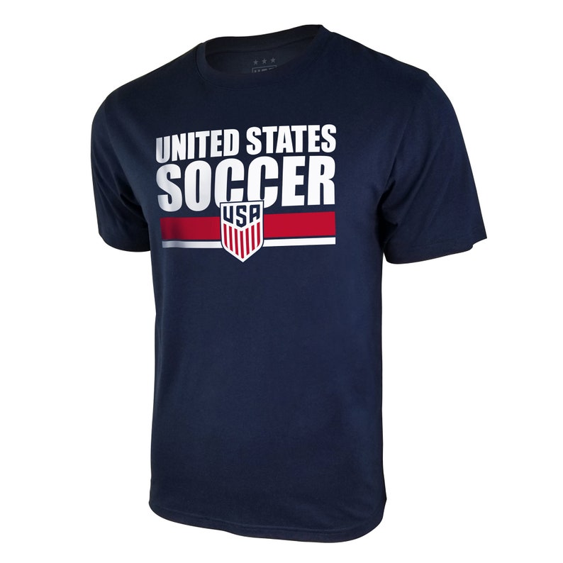 Pu&ograve; includere: Una maglietta blu navy con una grafica bianca che dice "UNITED STATES SOCCER" con uno scudo rosso, bianco e blu che dice "USA" sotto.