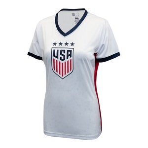 Puede incluir: Camiseta de f&uacute;tbol blanca con un escudo de EE. UU. azul y rojo y cinco estrellas sobre el escudo. La camiseta tiene un cuello y mangas azul marino con ribetes rojos en los lados.