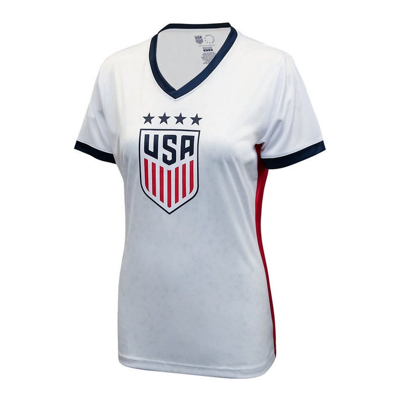 Puede incluir: Camiseta de f&uacute;tbol blanca con un escudo de EE. UU. azul y rojo y cinco estrellas sobre el escudo. La camiseta tiene un cuello y mangas azul marino con ribetes rojos en los lados.