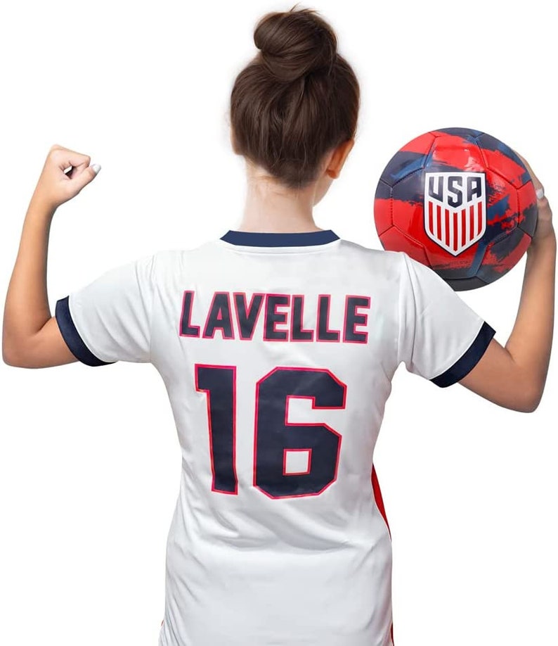 Op de afbeelding: Een wit voetbaltenue met blauwe bies en de naam "Lavelle" en nummer "16" op de rug. Het tenue wordt gedragen door een persoon met een knot. Een rood, wit en blauwe voetbal met het woord "USA" erop wordt in de andere hand gehouden.