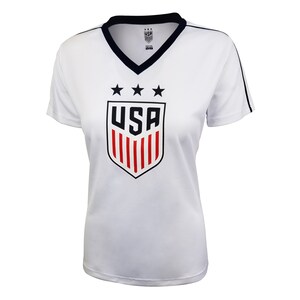 May include: White short-sleeved t-shirt with a navy blue v-neck collar and navy blue trim on the sleeves. The shirt features a red, white, and blue USA crest with three stars above the crest.
