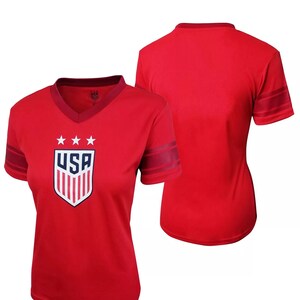 Puede incluir: Camiseta roja de manga corta con un logotipo de EE. UU. blanco y azul y rayas rojas y blancas en las mangas. La parte posterior de la camiseta es de color rojo liso.