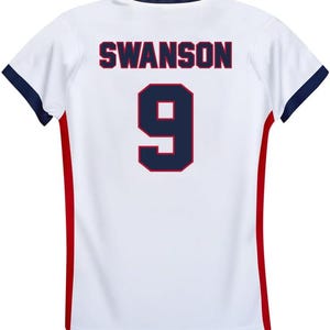 May include: White soccer jersey with red and blue trim. The back of the jersey has the name "SWANSON" and the number "9" printed in blue with a red outline.