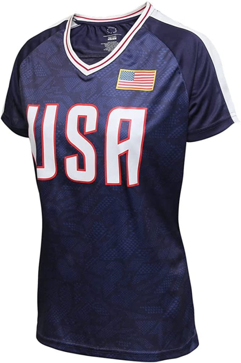 May include: Navy blue athletic shirt with the word "USA" in large white letters outlined in red. The shirt has a V-neck collar with red and white trim, white stripes on the sleeves, and an American flag patch.