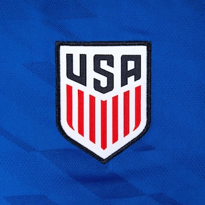 May include: Close-up of a blue athletic jersey with a USA emblem. The emblem is a white shield with the word "USA" in black block letters at the top and red vertical stripes at the bottom. The jersey has a textured pattern.