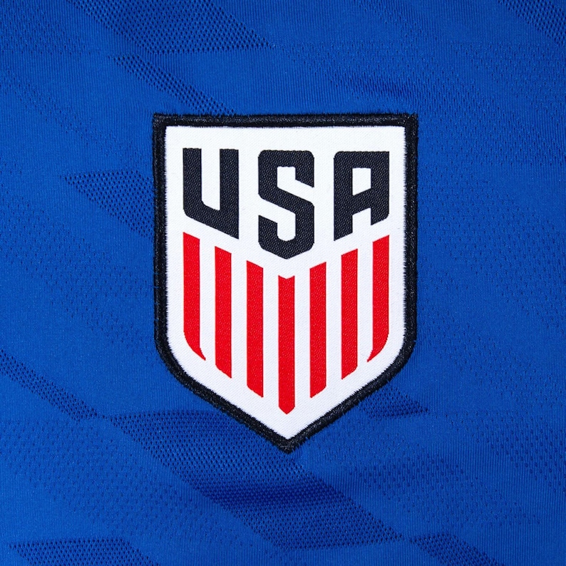 May include: Close-up of a blue athletic jersey with a USA emblem. The emblem is a white shield with the word "USA" in black block letters at the top and red vertical stripes at the bottom. The jersey has a textured pattern.
