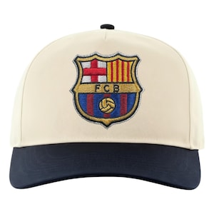 May include: A cream-colored baseball cap with a navy blue brim. The cap features an embroidered emblem of the FC Barcelona crest, including a red cross, yellow and red stripes, and a soccer ball.