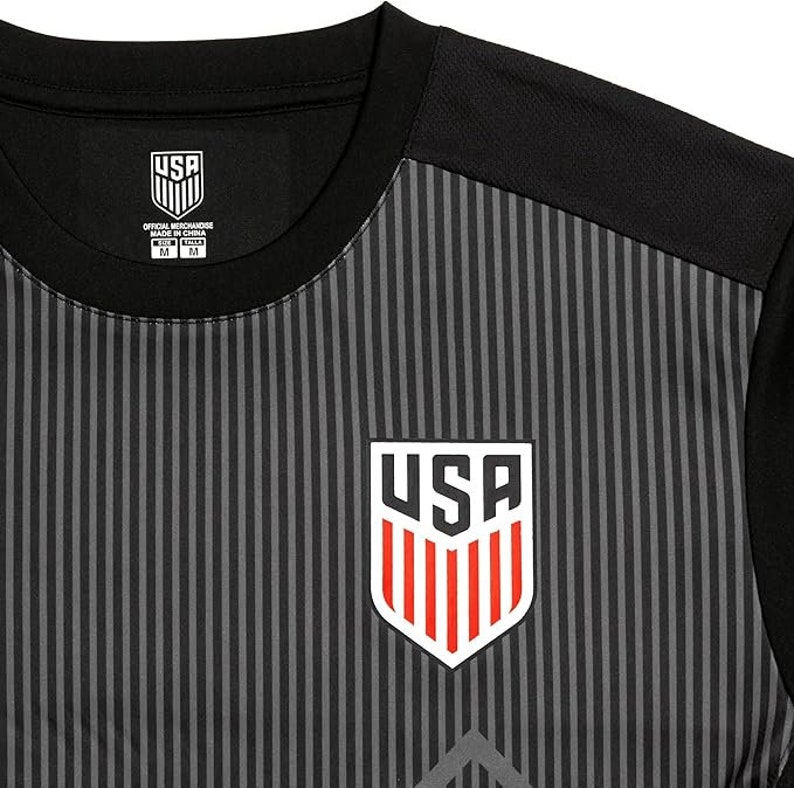May include: Black and gray striped soccer jersey with a white and red USA logo on the front. The jersey has a black collar and short sleeves. The tag on the jersey says "Official Merchandise Made in China".