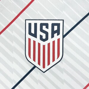 May include: Close-up of a white background with a shield-shaped emblem. The emblem features the letters "USA" in navy blue, above red vertical stripes. Diagonal red and navy blue lines intersect the background.