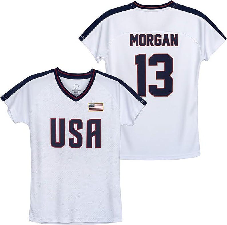 May include: White athletic jersey with navy blue accents. The front displays "USA" in navy lettering, a small American flag, and a V-neck collar. The back features "MORGAN" and the number "13" in navy.