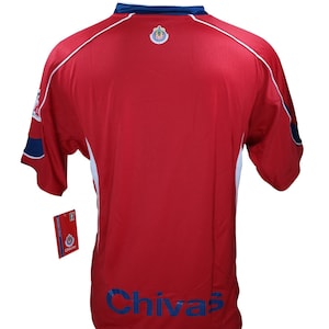 May include: Red soccer jersey with white and blue trim. The jersey has the team logo on the back and the word "Chivas" printed in blue on the bottom.