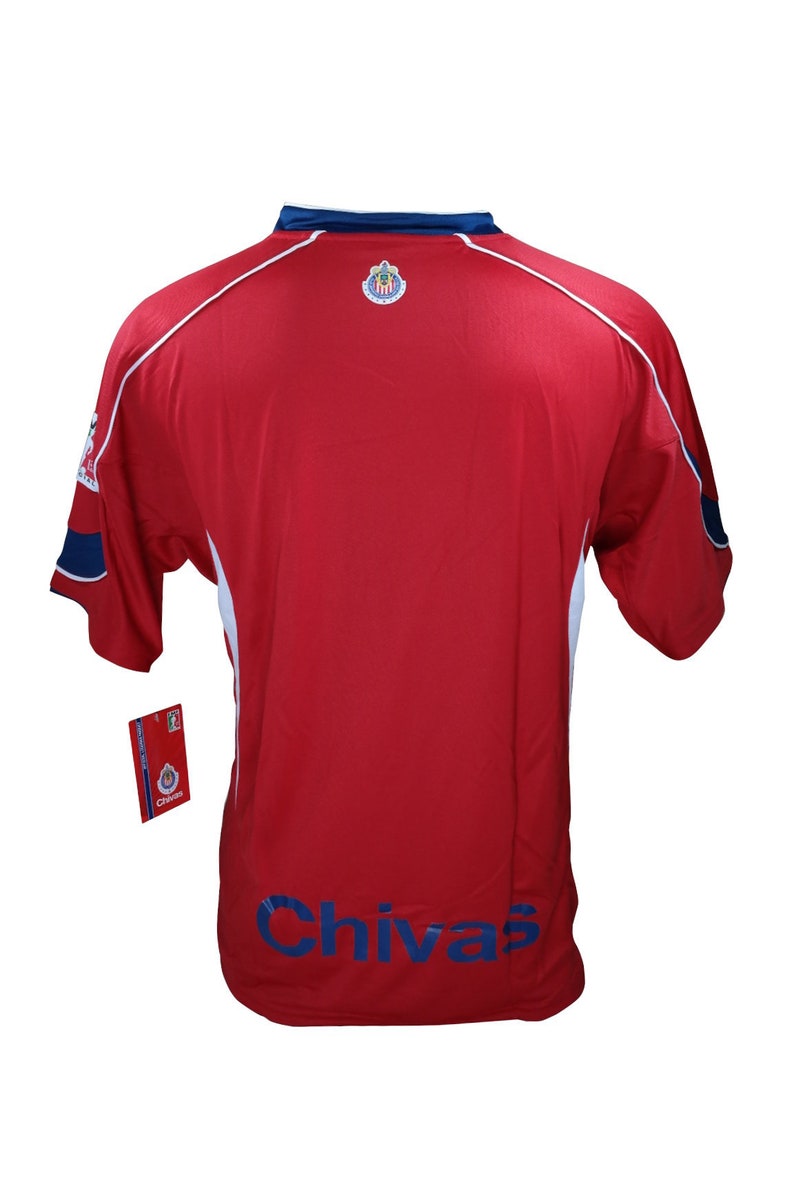 May include: Red soccer jersey with white and blue trim. The jersey has the team logo on the back and the word "Chivas" printed in blue on the bottom.