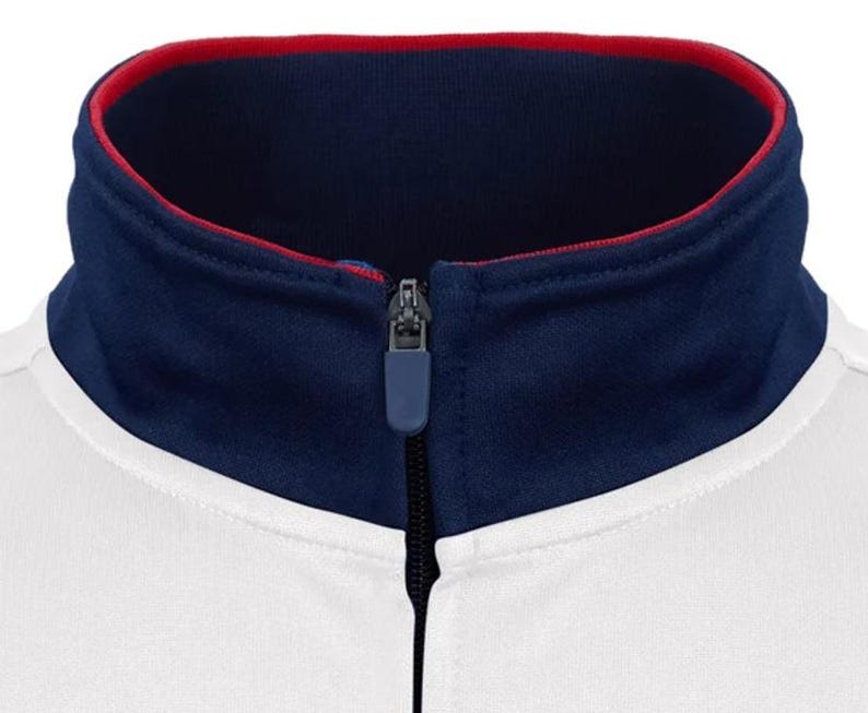 Puede incluir: Una chaqueta blanca con cremallera, con un cuello azul marino y ribete rojo. El cuello est&aacute; forrado con tela roja.