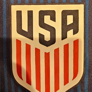 May include: A white patch with a black outline and the word "USA" in black letters. The patch has red and white stripes underneath the word "USA".