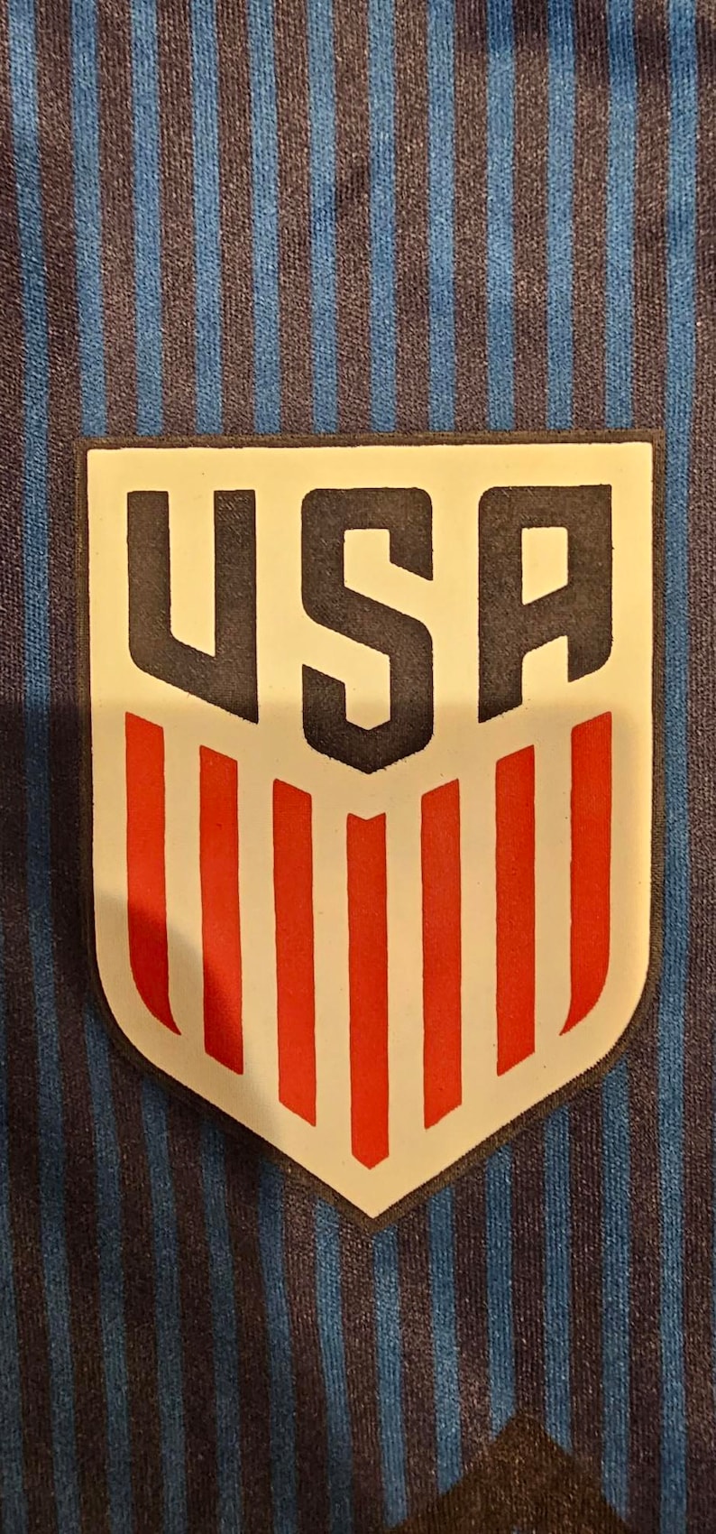 May include: A white patch with a black outline and the word "USA" in black letters. The patch has red and white stripes underneath the word "USA".
