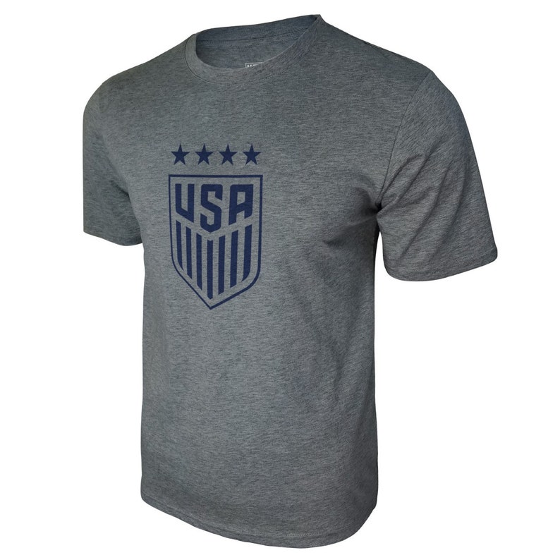 Puede incluir: Una camiseta gris jaspeada con un escudo de los Estados Unidos de color azul marino con cinco estrellas y rayas verticales. El escudo est&aacute; en la parte delantera de la camiseta.