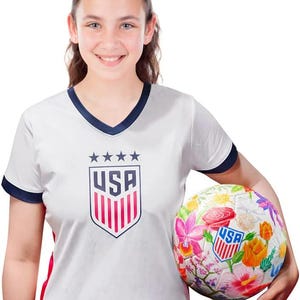 Puede incluir: Una camiseta de f&uacute;tbol blanca con cuello azul marino y rayas rojas y blancas en las mangas. La camiseta tiene un escudo blanco con la palabra "USA" y cinco estrellas. Una persona est&aacute; sosteniendo un bal&oacute;n de f&uacute;tbol blanco con un dise&ntilde;o floral y la palabra "USA" en &eacute;l.