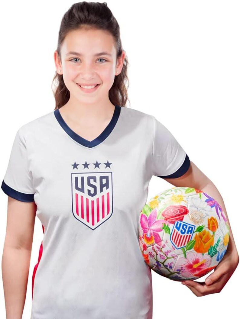 Puede incluir: Una camiseta de f&uacute;tbol blanca con cuello azul marino y rayas rojas y blancas en las mangas. La camiseta tiene un escudo blanco con la palabra "USA" y cinco estrellas. Una persona est&aacute; sosteniendo un bal&oacute;n de f&uacute;tbol blanco con un dise&ntilde;o floral y la palabra "USA" en &eacute;l.