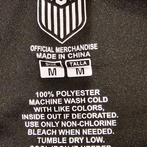 May include: A black tag with white text that reads "USA" in a bold font. The tag also includes care instructions for a 100% polyester garment. The tag states "Official Merchandise Made in China" and includes size information: "Size M Talla M".