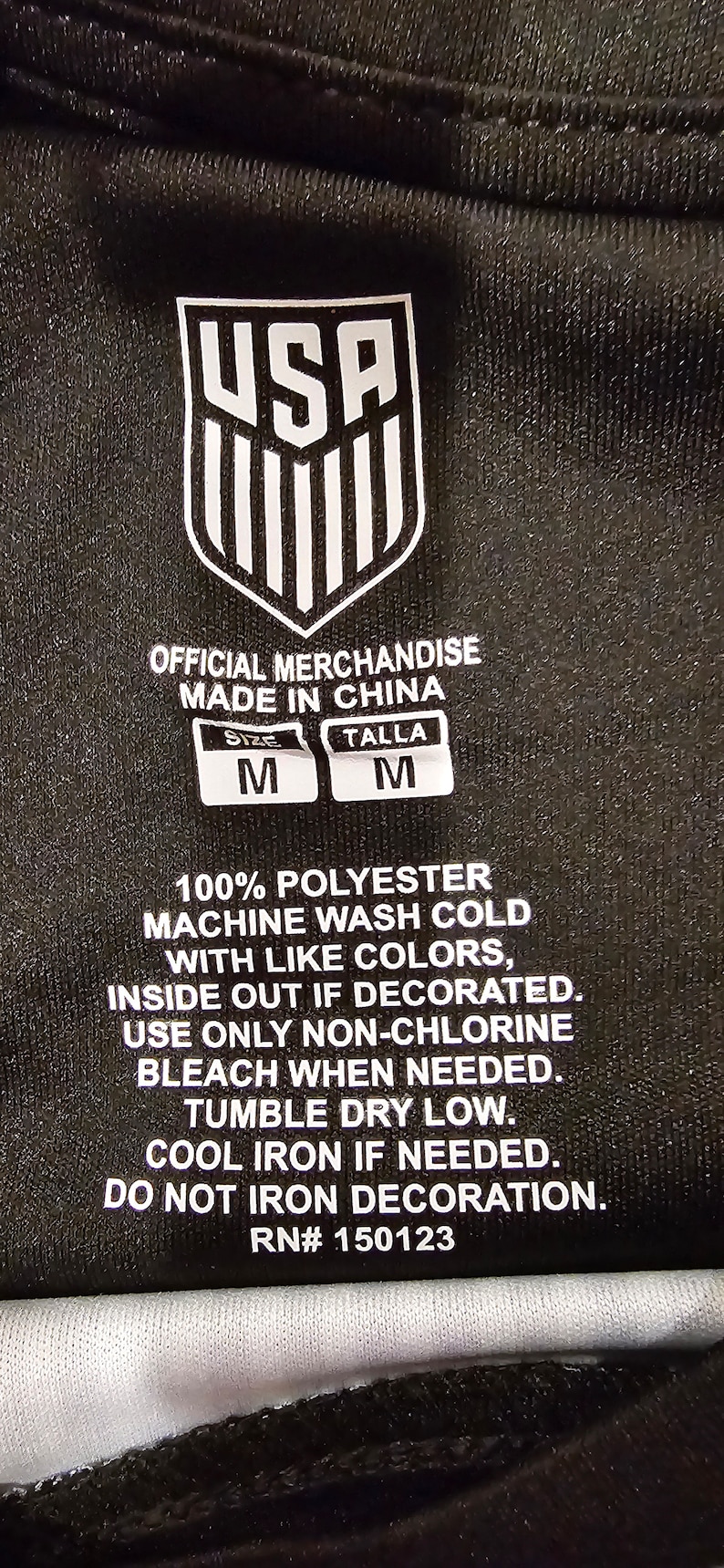 May include: A black tag with white text that reads "USA" in a bold font. The tag also includes care instructions for a 100% polyester garment. The tag states "Official Merchandise Made in China" and includes size information: "Size M Talla M".