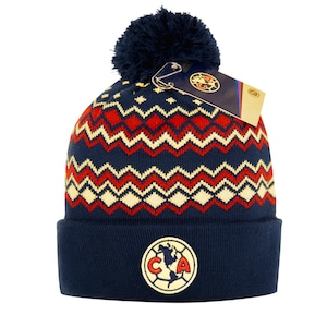 May include: A navy blue knitted beanie hat with a red, cream, and navy zigzag pattern. The hat has a navy pom-pom and a circular emblem with the letters "CA" and a map. A tag is attached.