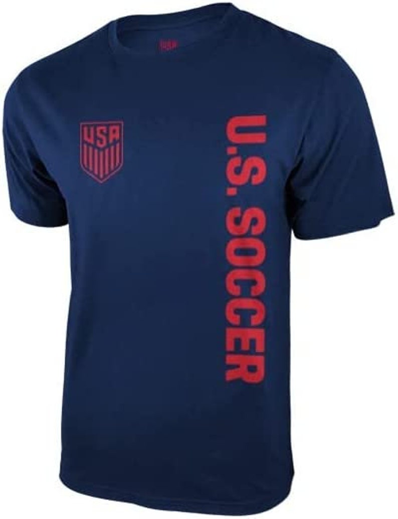 Puede incluir: Una camiseta azul marino con un gr&aacute;fico rojo "U.S. SOCCER" en el lado derecho y un logotipo rojo "USA" en el lado izquierdo.