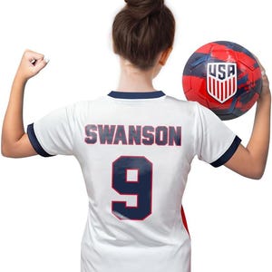 May include: White soccer jersey with blue trim and the name "SWANSON" and the number "9" printed on the back. The jersey is being held by a person holding a red, white, and blue soccer ball with the word "USA" printed on it.