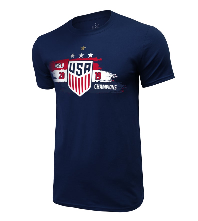 May include: Navy blue t-shirt with a white, red, and blue graphic of the USA soccer team logo. The graphic includes the text "WORLD 2019 CHAMPIONS" and three gold stars.