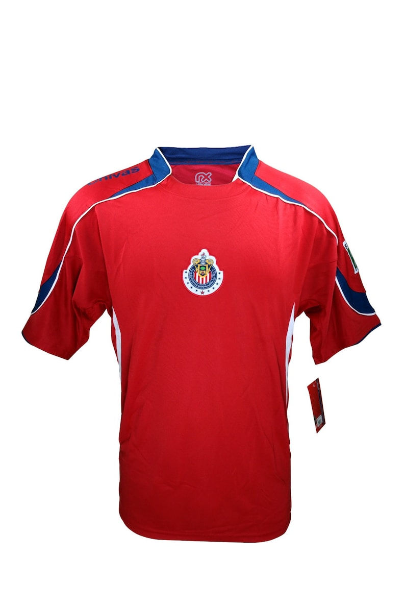 May include: Red soccer jersey with white and blue trim. The jersey features a team logo with a shield and a crown. The logo is in the center of the jersey.