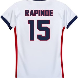 May include: White soccer jersey with red and blue trim. The back of the jersey has the name "RAPINOE" and the number "15" printed in navy blue.