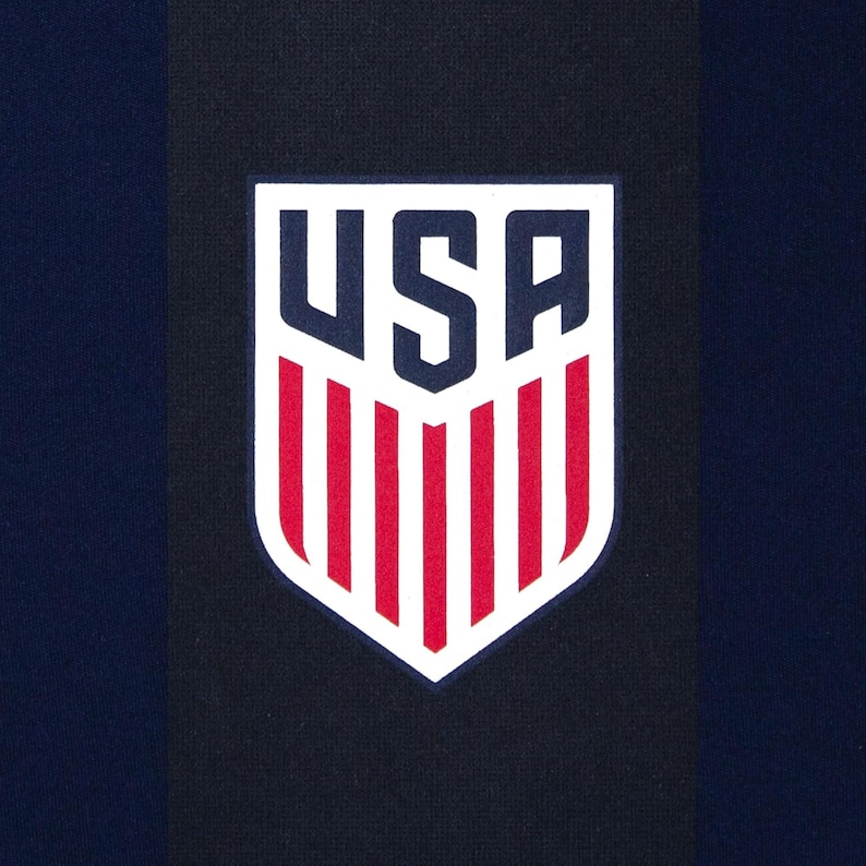 May include: Close-up of a navy blue fabric with a vertical black stripe. A white shield-shaped emblem with the letters "USA" in navy blue and red vertical stripes is centered on the fabric.
