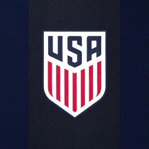 Może przedstawiać: Zbliżenie granatowej tkaniny z pionowym czarnym paskiem. Na tkaninie umieszczono białą tarczę z napisem "USA" w kolorze granatowym i czerwonymi pionowymi paskami.
