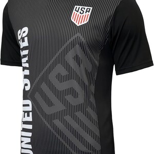 Puede incluir: Camiseta de f&uacute;tbol de manga corta negra con un dise&ntilde;o gr&aacute;fico gris y blanco. El gr&aacute;fico presenta las palabras "USA" y "UNITED STATES" y un escudo con las letras "USA" en su interior.
