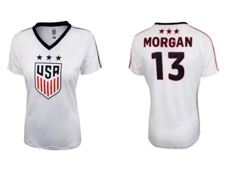 May include: White soccer jersey with blue and red accents. The front of the jersey features a USA shield logo with three stars above it. The back of the jersey has the name "MORGAN" and the number "13" printed in black with red outlines.