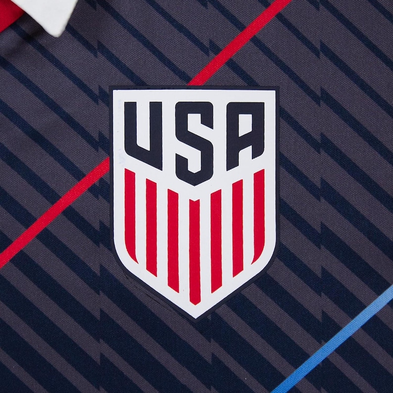 Peut inclure: Gros plan sur un maillot bleu marine et gris &agrave; motifs avec un col blanc. Le maillot pr&eacute;sente un &eacute;cusson blanc en forme de bouclier avec les lettres "USA" en noir et des rayures verticales rouges. Des rayures diagonales rouges et bleues accentuent le design.