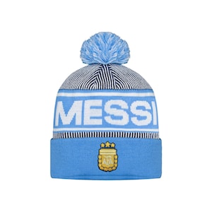 May include: A light blue knitted beanie hat with a matching pom-pom. The word "MESS" is displayed in white block letters across the front. Below the text is a light blue band with the Argentina Football Association logo.
